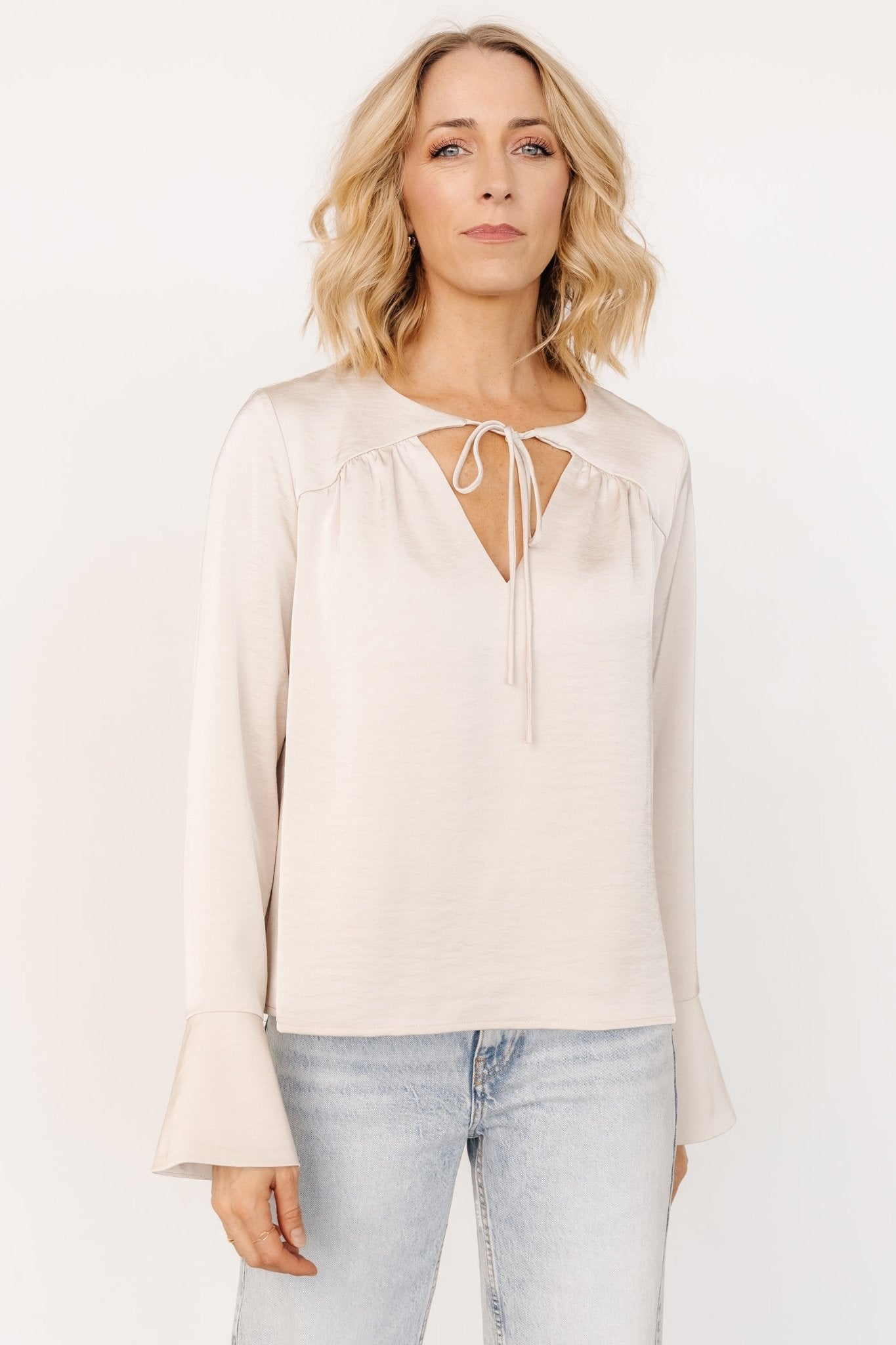 Front view of pearl champagne satin-textured blouse with V tie-neck, pleated front and long bell sleeves on model, draped fit.