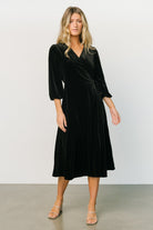 Close three-quarter view of black velvet wrap dress highlighting V-neck, wrap tie secured at side loop and soft gathered elastic cuffs.