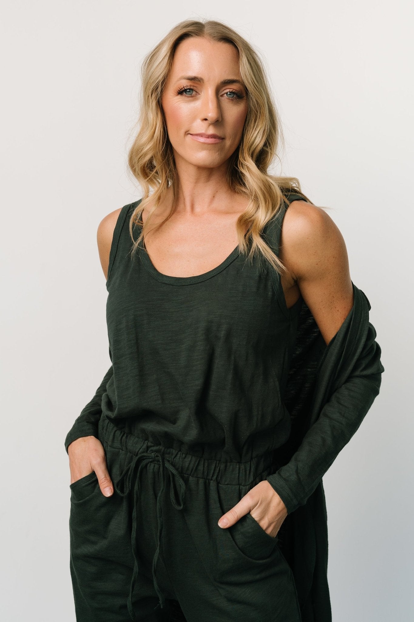 Side-angle of dark green jumpsuit and long cardigan on model, highlighting comfortable oversized fit, jogger cuffs, and functional side pockets.