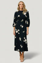 Styled shot of Karalie maxi dress on model, navy floral print, smocked waist, ruffled mock neck and mid-length sleeves visible.