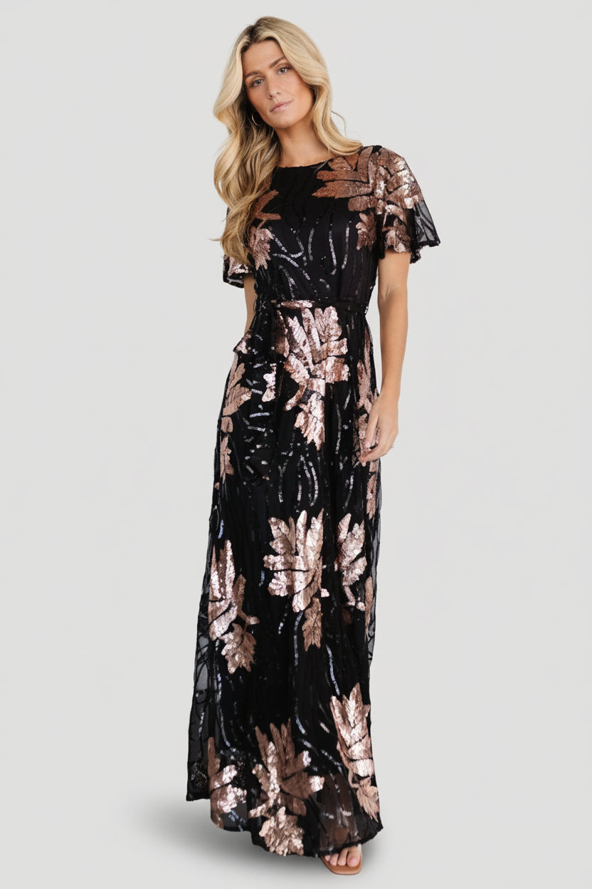 Front view of black maxi dress densely embroidered with rose-gold sequin leaf pattern, round neckline and tied waist sash for shape.