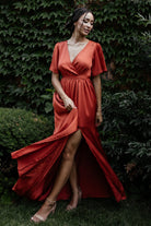 Full-length front view of rust satin-like maxi dress with surplice V-neck, flutter sleeves, elastic waist and removable sash worn by a model.