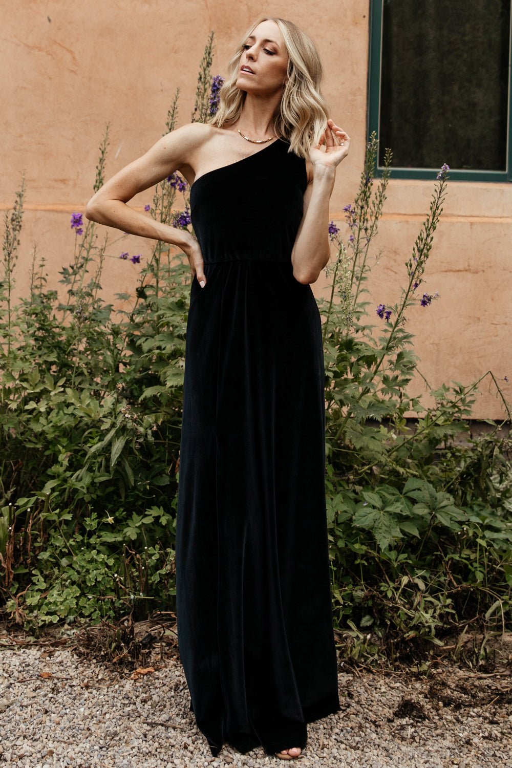 Model wearing black velvet one-shoulder maxi dress, front view showing fitted bodice, asymmetrical neckline, and slight skirt flare.