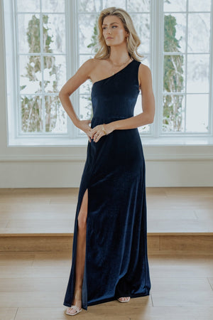 Front view of Tatiana navy velvet one-shoulder maxi dress on model showing fitted bodice, asymmetrical neckline, slight flare and right-side slit.